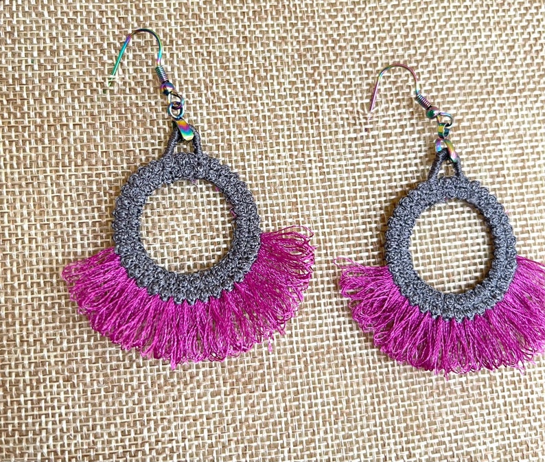 May include: A pair of gray crocheted hoop earrings with a pink fringe. The earrings have a simple, elegant design.
