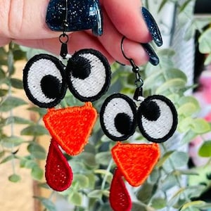 May include: A pair of turkey-themed earrings with red, orange, white, and black embroidered details. The earrings are made of felt and have a dangling design.