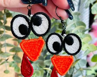 May include: A pair of turkey-themed earrings with red, orange, white, and black embroidered details. The earrings are made of felt and have a dangling design.