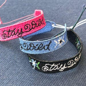 May include: Three embroidered bracelets with the phrase "Stay Wild" in black lettering. The bracelets are pink, blue, and black.