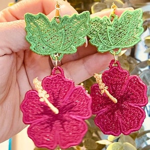 May include: A pair of dangle earrings featuring a green leafy design and a pink hibiscus flower. The earrings are made of embroidered fabric and have gold-colored hooks.