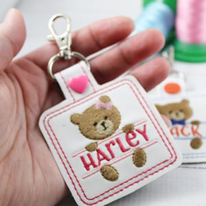 May include: White fabric keychain with a pink stitched border. The keychain features a brown teddy bear with a pink bow and the name "Harley" stitched in pink. A pink heart charm is attached to the keychain.