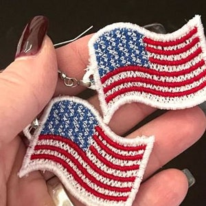 May include: A pair of earrings featuring a red, white, and blue American flag design. The flags are embroidered and have a wavy, flowing shape.