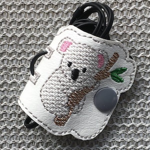May include: White leather cord keeper with an embroidered koala design. The koala is gray with brown branches and green leaves. The cord keeper has a gray button.