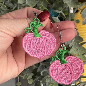 以下が含まれることがあります： 緑色の茎が付いたピンク色の刺繍カボチャのイヤリング。これらのイヤリングは、秋やハロウィーンに最適なお祝いのアクセサリーです。カボチャのデザインは、質感のあるステッチの外観で詳細に表現されています。