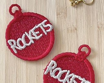 Pode incluir: Brincos vermelhos bordados com a palavra "ROCKETS" em branco. Os brincos são redondos com uma argola no topo e ganchos dourados.