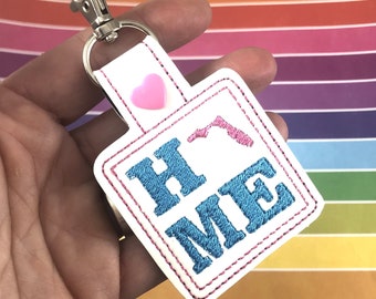 May include: White rectangular keychain with the word "HOME" embroidered in blue, outlined in pink. A pink heart and a pink state outline are also present. The keychain has a silver clasp.