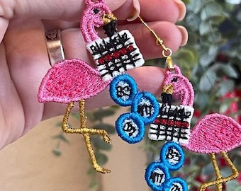 May include: Embroidered flamingo earrings with gold-colored hooks. The pink flamingos have gold legs and are paired with blue and white embroidered accents. The earrings have a unique, handmade appearance.