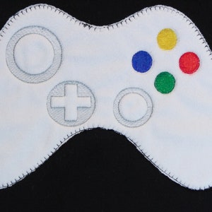 May include: White fabric game controller with grey buttons and a black stitched border. The controller has four coloured buttons: yellow, blue, red and green.