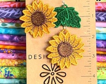 Puede incluir: Un par de pendientes colgantes de girasol y hoja verde. Los pendientes se muestran en una tarjeta marrón con el texto "DESIGNS by babymoon".