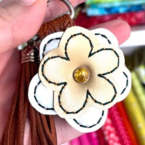May include: A gold and white leather flower keyring with a brown tassel. The flower has a green stitched outline and a yellow rhinestone in the centre.