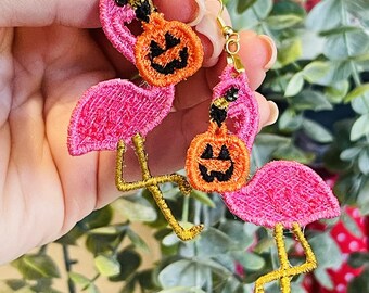 May include: Pink flamingo earrings with gold-colored legs and hooks. Each flamingo holds an orange pumpkin with a carved face. The earrings are made of embroidered fabric, perfect for Halloween.