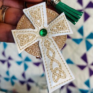 May include: A Celtic cross keyring with a gold glitter background and a white cross overlay. The cross features intricate beige embroidery and a green jewel centre. A green tassel hangs from the top.
