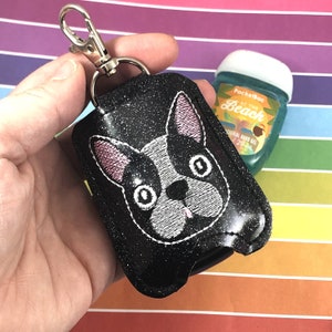 May include: Black glitter hand sanitizer holder with a white and pink embroidered French bulldog design. The holder has a silver keychain attached.