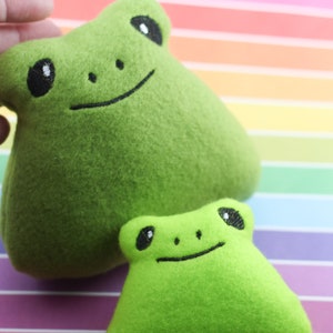 Puede incluir: Dos juguetes de peluche de rana verde con ojos y sonrisas bordados. La rana más grande está en primer plano y la rana más pequeña está en el fondo.