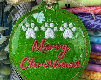 May include: A green Christmas ornament with white paw prints and the embroidered text "Merry Christmas" in red. The ornament has a gold ribbon and a red bead for hanging. The background shows colorful fabric.