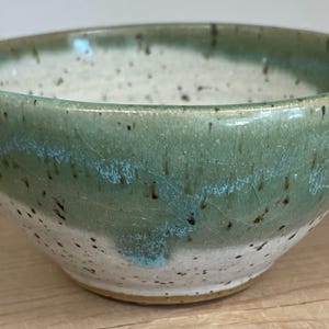 May include: A small, white and green ceramic bowl with a speckled glaze. The bowl has a rounded shape and a slightly flared rim.