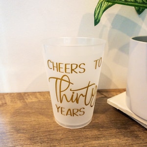 K&ouml;nnte beinhalten: Ein gefrosteter Plastikbecher mit dem Text "Cheers to Thirty Years" in goldenen Buchstaben.