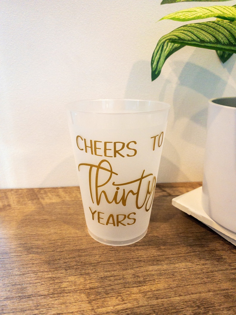 K&ouml;nnte beinhalten: Ein gefrosteter Plastikbecher mit dem Text "Cheers to Thirty Years" in goldenen Buchstaben.