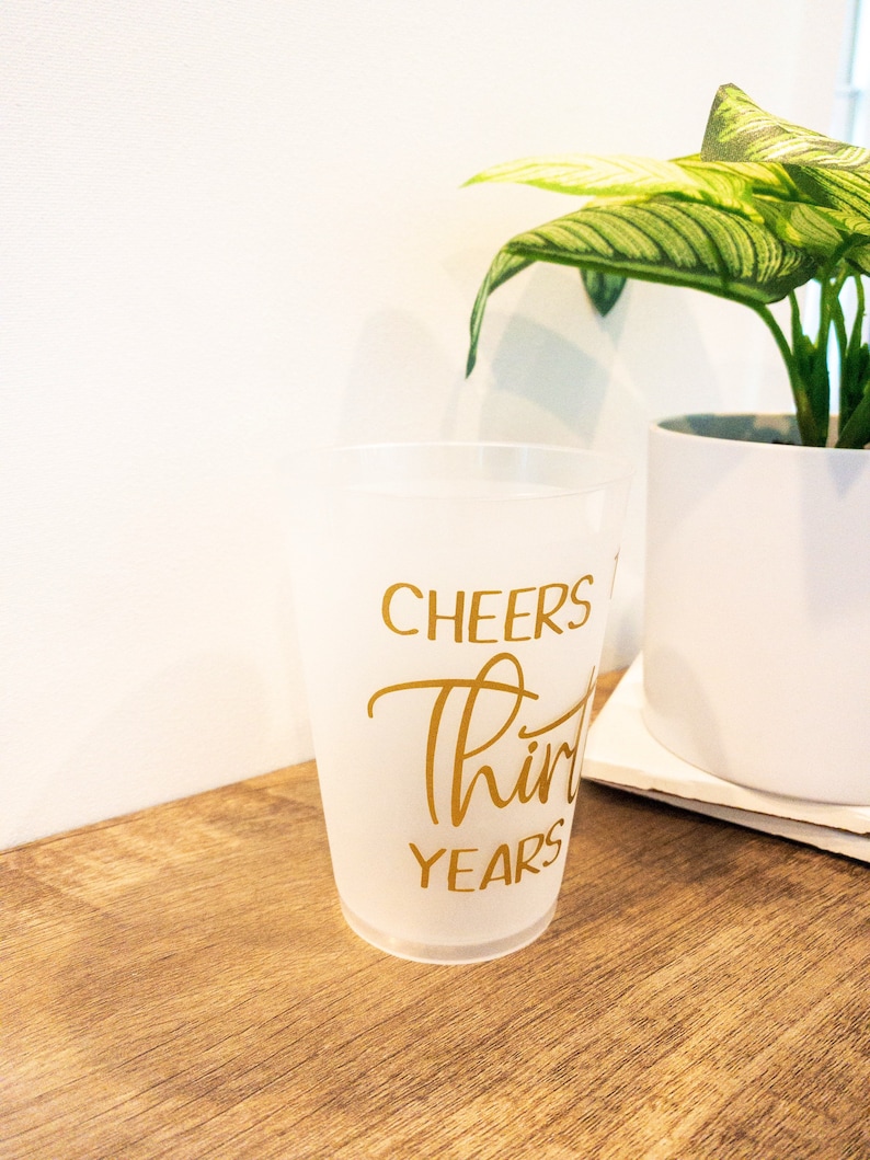 K&ouml;nnte beinhalten: Ein gefrosteter Plastikbecher mit dem Text "Cheers to Thirty Years" in goldenen Buchstaben.