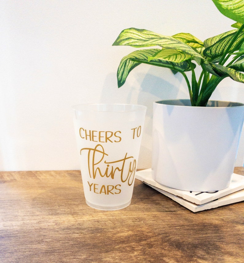 K&ouml;nnte beinhalten: Ein durchsichtiger Plastikbecher mit dem Aufdruck "Cheers to Thirty Years" in goldener Schrift.