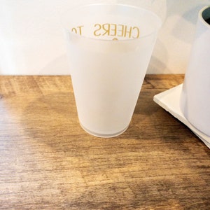 K&ouml;nnte beinhalten: Ein wei&szlig;er, gefrosteter Plastikbecher mit dem Text "CHEERS TO" in Gold auf der Seite.