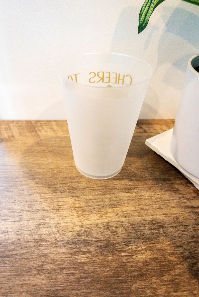 K&ouml;nnte beinhalten: Ein wei&szlig;er, gefrosteter Plastikbecher mit dem Text "CHEERS TO" in Gold auf der Seite.
