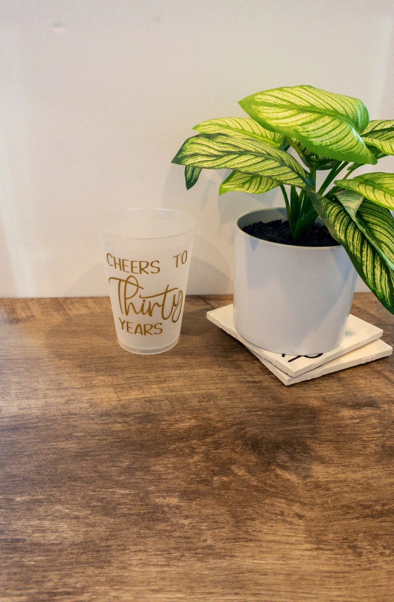 K&ouml;nnte beinhalten: Ein durchsichtiger Plastikbecher mit dem Aufdruck "Cheers to Thirty Years" in goldenen Buchstaben. Der Becher steht auf einem Holztisch neben einer Topfpflanze mit gr&uuml;n-wei&szlig;en Bl&auml;ttern.