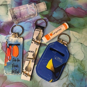 May include: Four keychains with different designs. One keychain has a blue background with a yellow fish and the text "Just Keep Swimming". Another keychain has a white background with a seagull and the text "Mine". A third keychain has a blue background with a fish and the text "Fish Are Friends, Not Food". The fourth keychain is a lip balm holder with a tangerine lip balm.