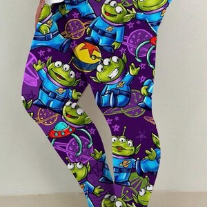 May include: Purple leggings featuring a repeating pattern of green, three-eyed aliens in blue space suits. The design includes planets, stars, and a yellow helmet. The leggings are worn by a person.