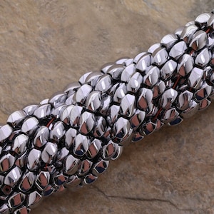May include: A close-up of a silver metal chain made of small, faceted beads. The chain is twisted and looped, showing the intricate design of the beads.