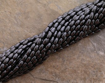 Peut inclure: Gros plan sur un fil de perles ovales gris foncé. Les perles sont enfilées ensemble, créant un motif texturé en forme de serpent. Le fond est un brun terreux moucheté.