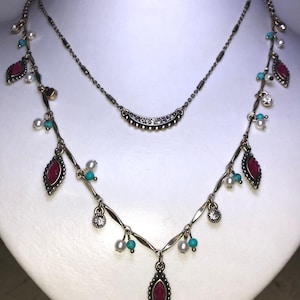 May include: Two-strand necklace with a vintage aesthetic. The lower strand features ruby-red teardrop-shaped pendants, turquoise beads, and small white pearls. The upper strand is a delicate chain with a curved, gem-studded design. The necklaces are displayed on a white bust.
