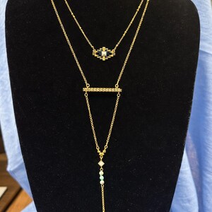 May include: A layered gold-toned necklace set. The top necklace features a geometric pendant with black and white accents. The lower necklace has a bar pendant with a vertical chain and small beads. The necklaces are displayed on a black mannequin.