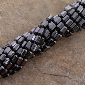 May include: A strand of black hematite beads in a faceted, rectangular shape. The beads are arranged in a spiral pattern and are displayed on a textured, brown surface.