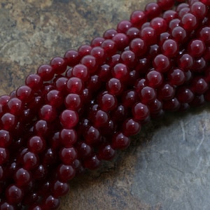 Puede incluir: Una hebra de cuentas de piedras preciosas de color rojo intenso. Las cuentas redondas son lisas y pulidas, creando un aspecto uniforme.