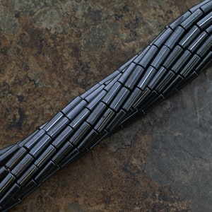 May include: A strand of black hematite beads, each bead is a cylinder with a slightly rounded edge. The beads are arranged in a spiral pattern on a brown textured surface.