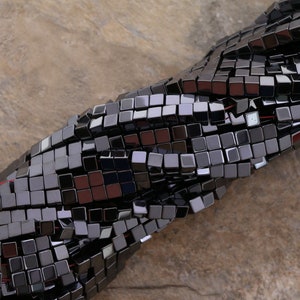 Puede incluir: Una hebra de cuentas de hematites negras, cada cuenta es un cubo con un acabado brillante y pulido. Las cuentas están enhebradas juntas en un patrón suelto y fluido.