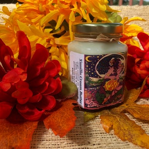 May include: A small glass jar with a gold lid filled with a white cream. The jar has a colorful label with the text "Vanilla Cream Butter" and a whimsical design featuring a woman and a crescent moon.