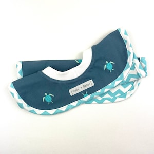 May include: A blue baby bib with a white trim and a turquoise chevron pattern. The bib features small turquoise turtle designs and a label that reads "Bella 'n Baker". The bib is folded and appears to be made of soft fabric.