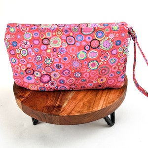 May include: A pink and red fabric pouch with a colorful circle pattern and a matching handle. The pouch is sitting on a wooden stand.