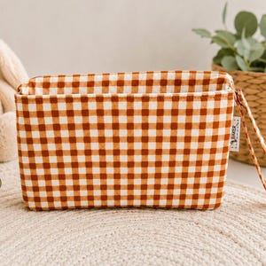 May include: A rectangular fabric pouch featuring a brown and white gingham check pattern. The pouch has a top zipper closure and a matching fabric wrist strap attached to the side. A small white tag with the text BAKER is sewn into the side seam.