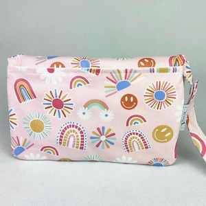 May include: A pink fabric pouch with a colorful pattern of rainbows, suns, flowers, and smiley faces. The pouch has a wrist strap and a zipper closure. The word "BAKER" is printed on the strap.