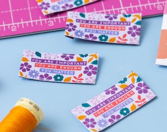 Könnte beinhalten: Vier weiße Stoff-Etiketten mit einem Blumenmuster und dem Text "You are important, You are enough, You matter" in lila, rosa und orangefarbener Schrift.