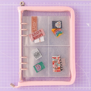 Peut inclure: Une pochette zippée rose avec des poches en plastique transparent. Les poches contiennent des patchs en tissu colorés avec différentes phrases, notamment "Made to Party", "Move More", "Love Yourself", "You Are Loved", "Stay True to You" et "Made by Auntie".