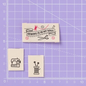 May include: Three fabric labels with sewing-themed designs. One label reads "SEWING IS IN MY SOUL" with a spool, scissors, and a button. The other two labels feature a sewing machine and a spool of thread with pins. All labels are on a purple grid.
