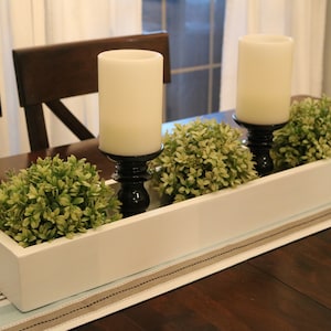 Puede incluir: Una jardinera de madera blanca con tres plantas verdes y dos velas blancas sobre candelabros negros. La jardinera está sobre una mesa de madera marrón con un camino de mesa azul y blanco.
