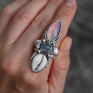 May include: A silver ring featuring a teardrop-shaped iridescent stone at the top, a small dark oval stone in the center, and a white cowrie shell at the base. The metal setting is detailed with small silver beads and decorative metal accents.