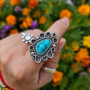 Op de afbeelding: Een zilveren ring met een turquoise steen in een decoratief ontwerp. De ring wordt gedragen op een vinger met meerdere armbanden.