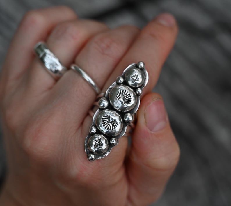 May include: A silver ring with an elongated design featuring multiple textured, circular elements. The ring has a series of smaller, round accents and is worn on a finger. Other silver rings are also visible.